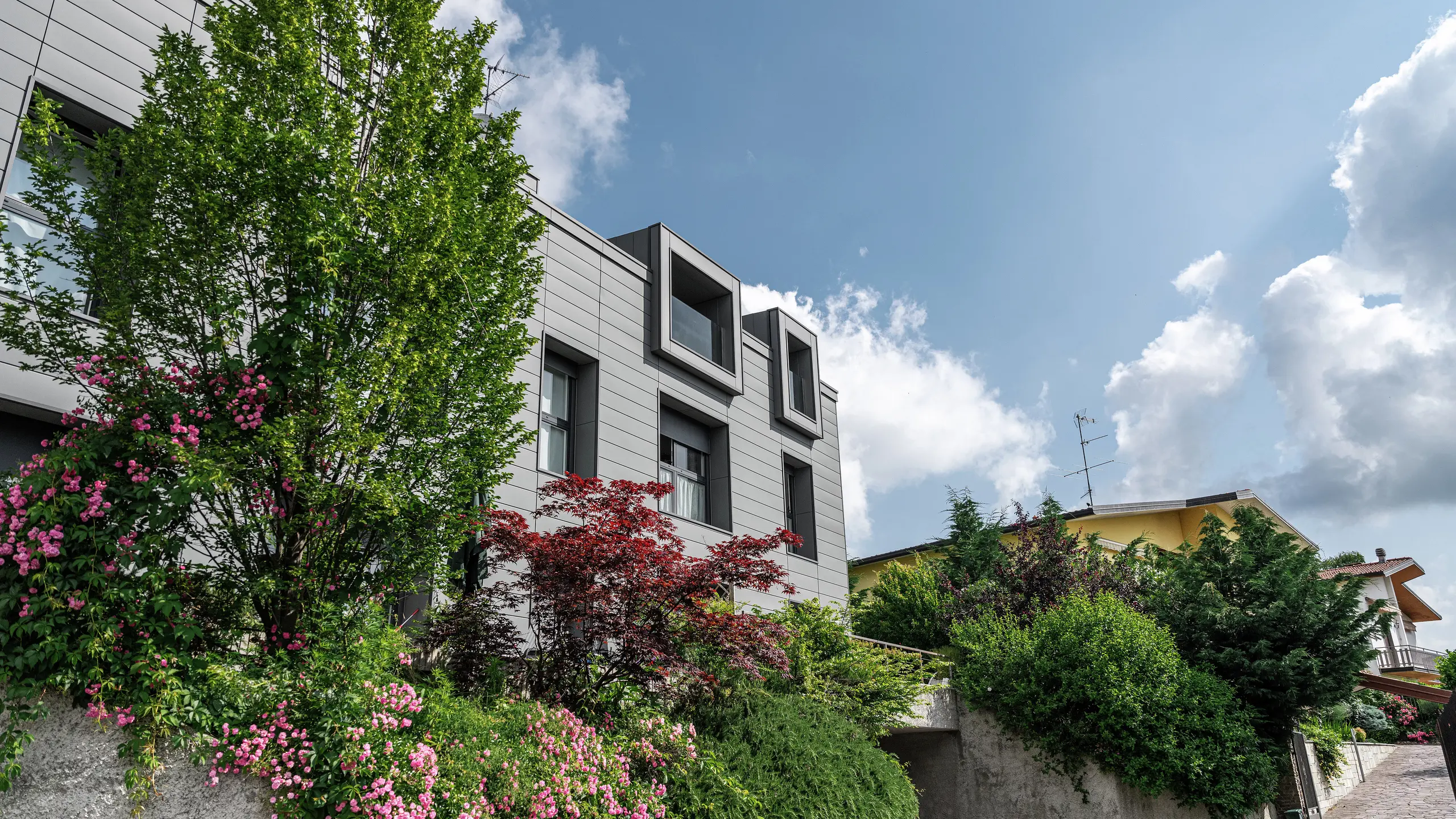 Modern wooncomplex in Faloppio, Italië, met stijlvolle gevel van PREFA Sidings. De uitkragende ramen vormen een unieke architectonische blikvanger, terwijl de omringende planten en bloemen de natuurlijke schoonheid van het object onderstrepen. De duurzame aluminium bekleding van PREFA zorgt voor een esthetische en functionele oplossing onder alle weersomstandigheden.