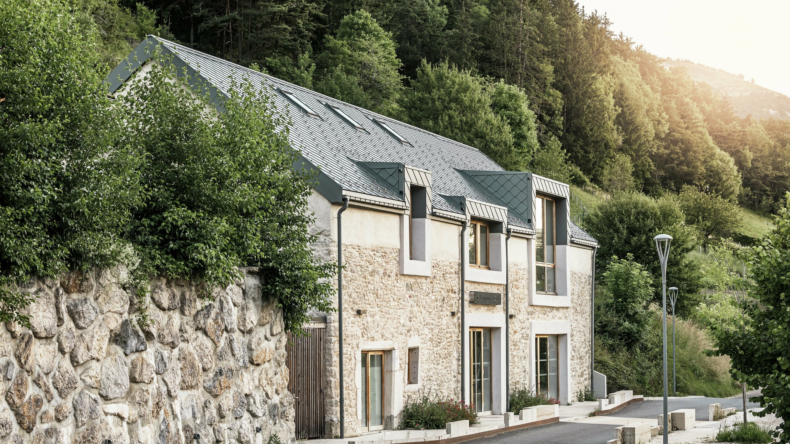 Seitenansicht der sanierten Scheune "La Grange à Gaby" in Engins, Frankreich. Das Gebäude kombiniert traditionelles Steinemauerwerk mit einem modernen PREFA Aluminiumdach aus 29 × 29 cm großen Rauten. Die behutsame Renovierung verbindet historische Bauweise mit langlebigen Materialien.