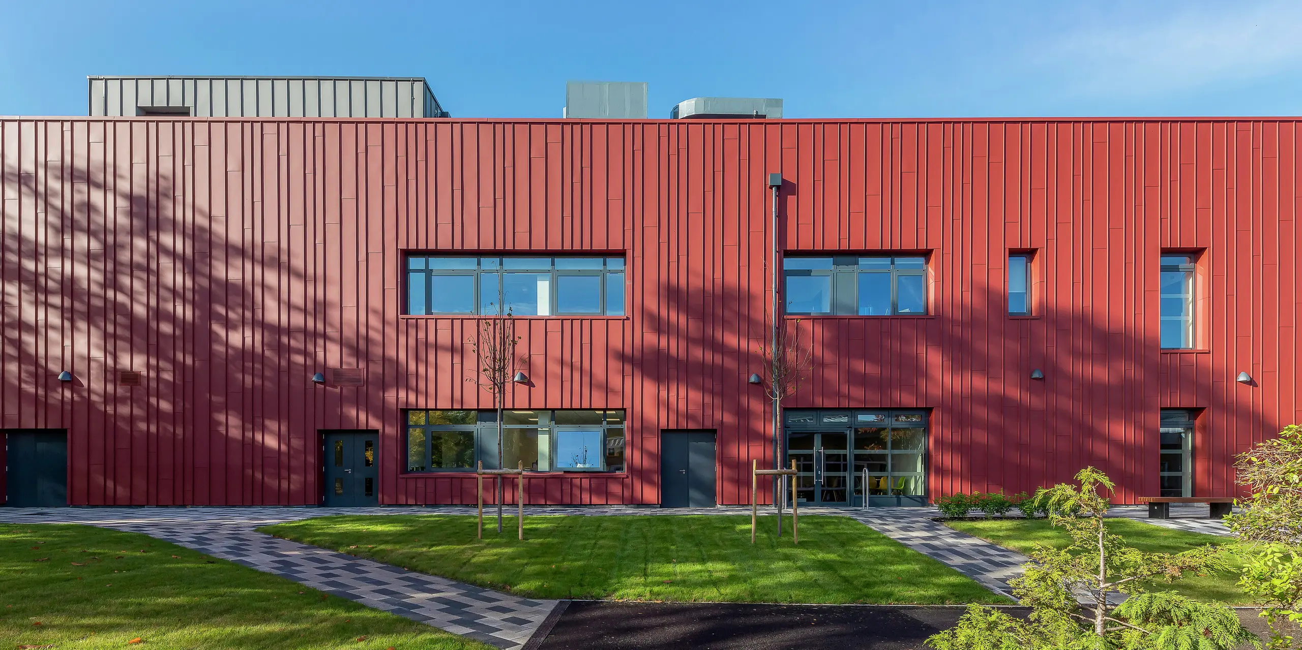 Moderne Fassadengestaltung mit PREFALZ: Das Soldiers' Centre in Pirbright besticht durch seine hochwertige Stehfalzverkleidung aus PREFALZ in P.10 Oxydrot. Die stilvolle Kombination mit Fensterbekleidungen in P.10 Anthrazit verleiht dem Sozialzentrum der Armee ein markantes Erscheinungsbild. Hier verbinden sich Funktionalität und Design - ein nachhaltiges Architekturkonzept mit langlebigem Aluminium.