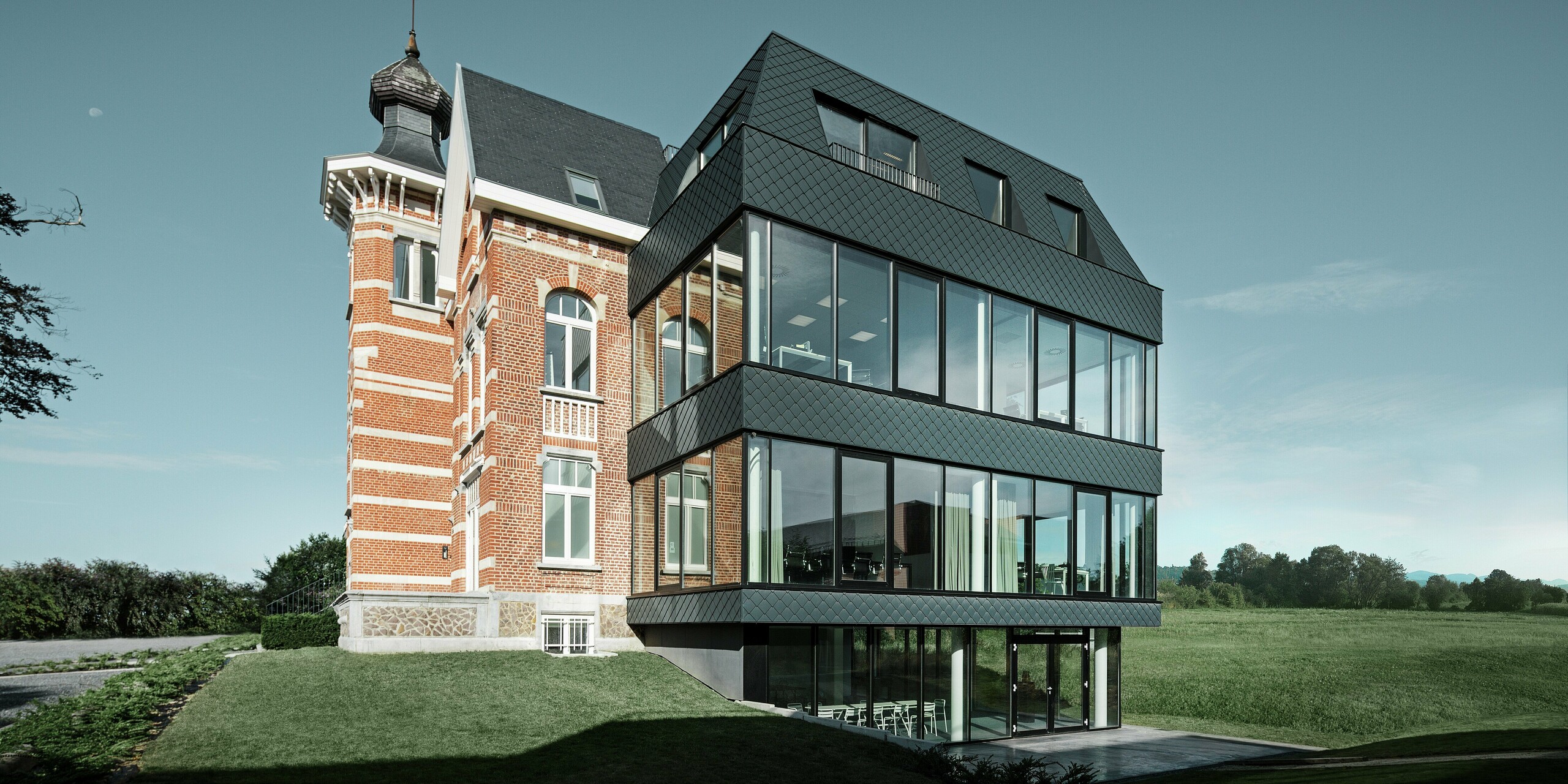 Seitlicher Blick auf das Kasteeltje Heizel, wo historischer Backsteinbau mit Turm nahtlos in einen modernen Anbau übergeht. Die anthrazitfarbenen PREFA Wandrauten setzen mit ihrer markanten Struktur elegante Kontraste zu den großzügigen Glasflächen. Die klare Linienführung des Neubaus verstärkt die Verbindung von Tradition und moderner Architektur in einer eindrucksvollen Kulisse.