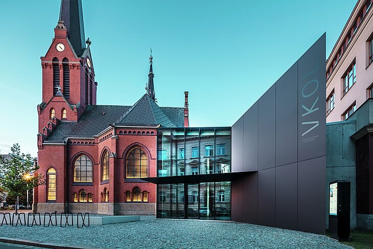 Das Bild zeigt das imposante Zusammenspiel zwischen der historischen Roten Kirche und dem modernen Neubau in Olmütz. Die Kirche mit ihren charakteristischen gotischen Fenstern und dem markanten Turm bildet das historische Rückgrat des Ensembles, während der moderne Anbau mit seiner eleganten, schwarzen PREFABOND Aluminium-Verbundplatten-Fassade eine zeitgemäße, architektonische Antwort darstellt. Große Glasflächen lassen den Blick in das Innere des Neubaus zu, wo die Umgebung durch Reflexionen in den Fenstern eine interessante Wechselwirkung zwischen Alt und Neu widerspiegelt. Die präzise Verarbeitung der PREFABOND-Platten betont die moderne Ästhetik, während die harmonische Integration in das historische Umfeld sowohl funktional als auch visuell beeindruckt.