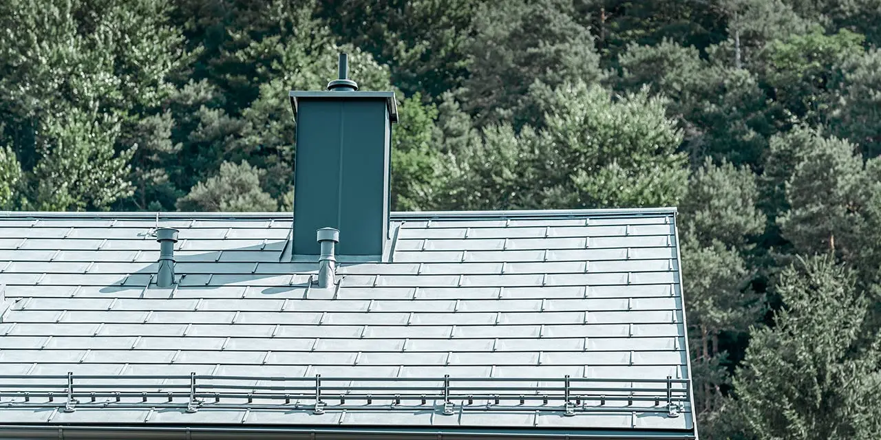Detailbeeld van een zadeldak, gedekt met de PREFA dakpan R.16 in steengrijs met sneeuwvangsysteem, schoornsteenomsluiting, dakgoot en ontluchtingspijp, eveneens van PREFA