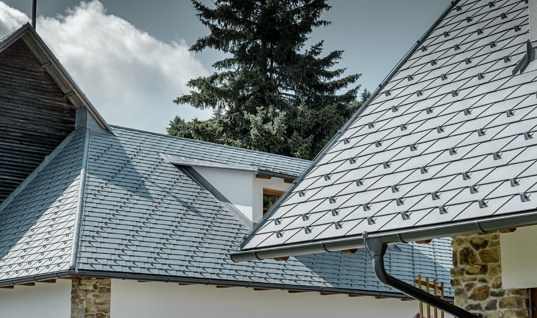 Detailopname van een aluminium dakbedekking van PREFA; dakschindel in steengrijs met de aluminium dakgoot in antraciet van PREFA; op de achtergrond herkent men een dakkapel met lessenaarsdak met staand felsdak. De gevel is wit met ingewerkte steenelementen.