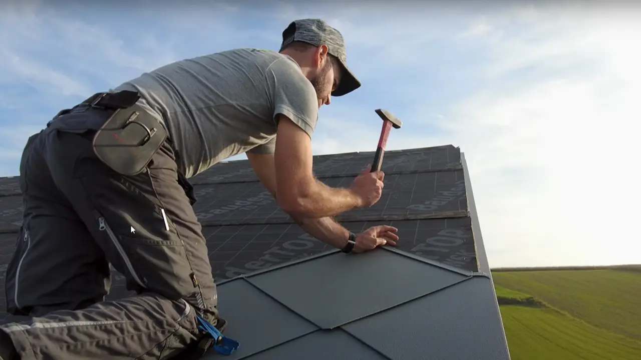 Loodgieter in actie: met een hamer legt de eigenaar van het bedrijf ‘Der Blechmann’ nauwkeurig de PREFA daklosange 44 × 44. De precisie bij de installatie is te zien aan de reeds gelegde daklosangen aan de onderkant van de foto. De man draagt een pet en knielt met zijn rug naar de camera op het dak. Op de achtergrond zijn de heldere lucht en een groen landschap te zien. De harmonieuze achtergrond onderstreept de symboliek van de foto: duurzaam en langdurig vakmanschap met hoogwaardige PREFA aluminiumproducten.