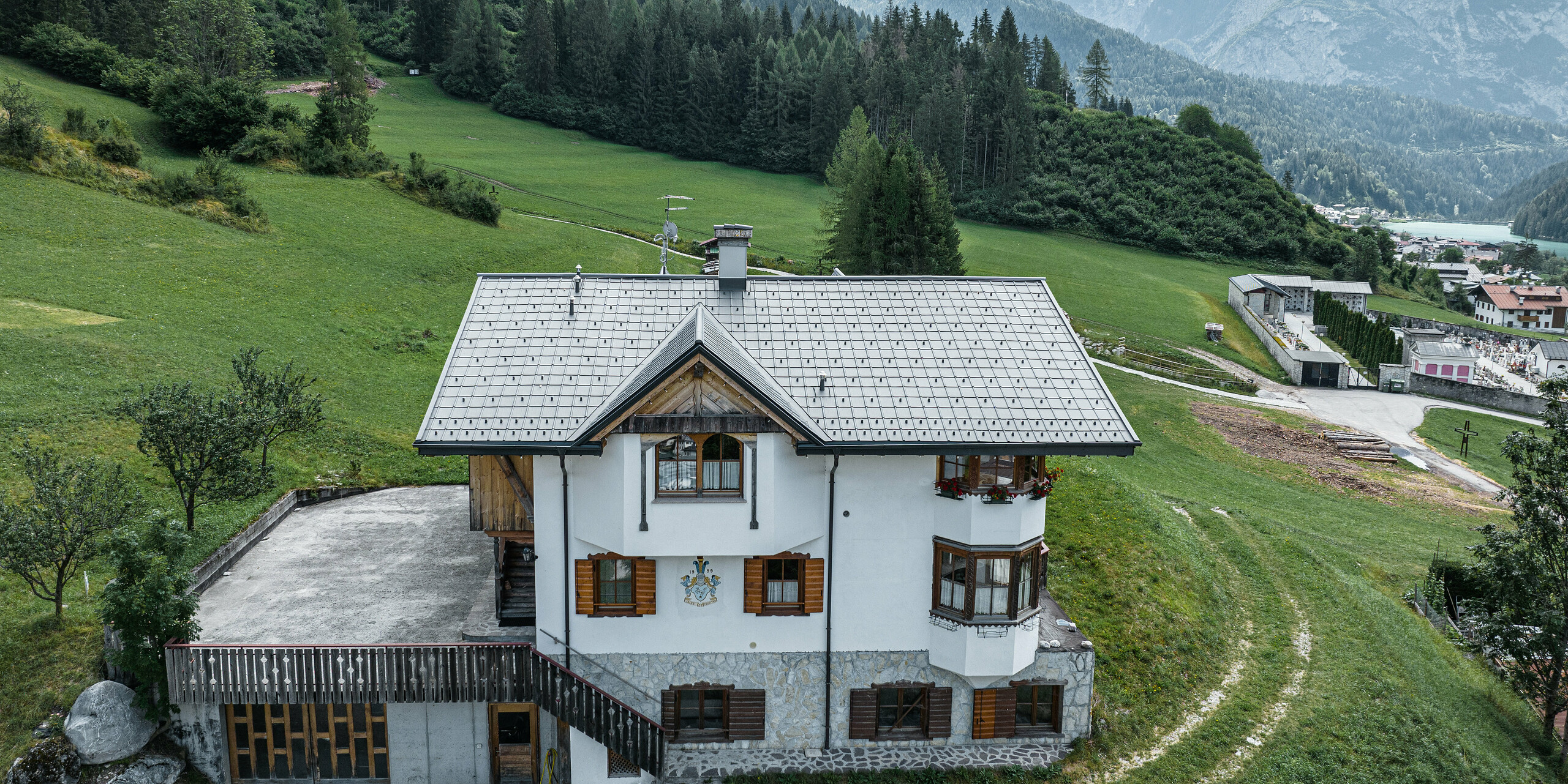 Vogelvluchtperspectief van de ingang van de traditionele berghut in Auronzo di Cadore na de dakrenovatie met PREFA aluminium dakpannen in P.10 lichtgrijs. Het gebouw met zijn karakteristieke alpine charme en het moderne tinnen dak past harmonisch in het weelderige groene landschap en het indrukwekkende bergpanorama op de achtergrond en toont een geslaagde combinatie van functionaliteit en regionale architectuur.
