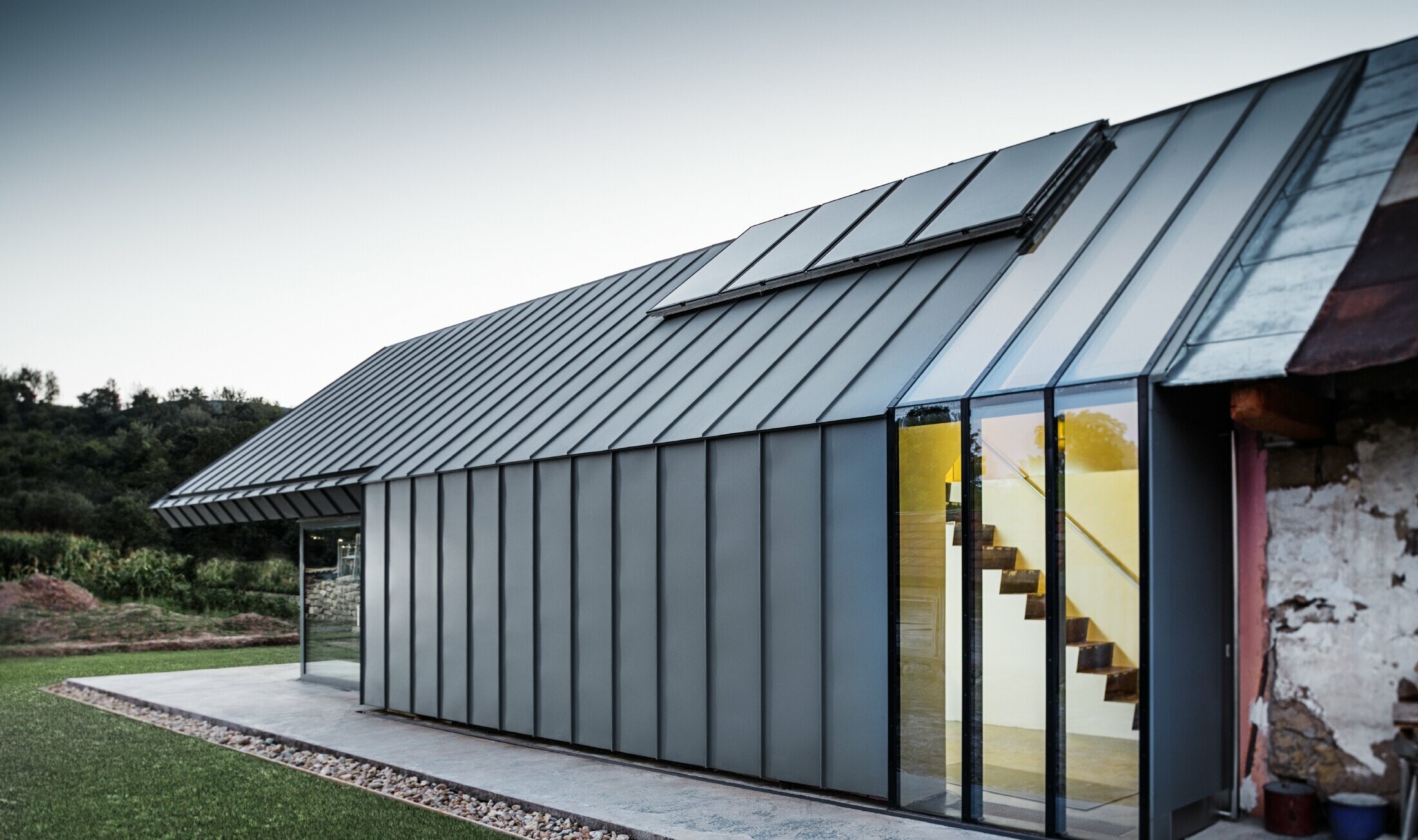 Modern architectenbureau/woning gebouwd tegen een oude boerderij, bekleed met staande naden en gebogen staande naden van PREFA in lichtgrijs