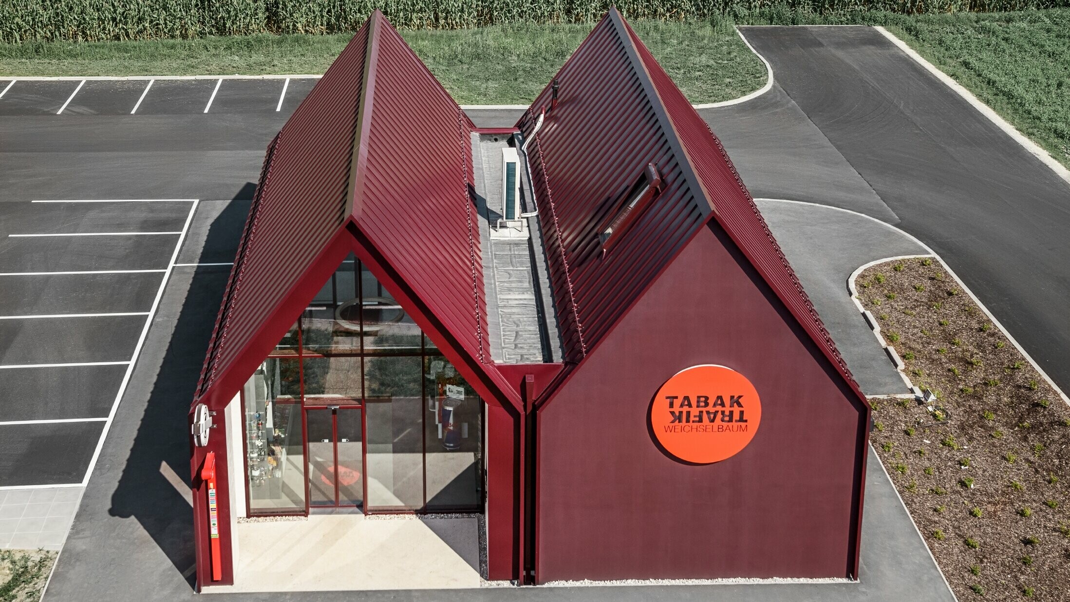Moderne tabakswinkel met zadeldak zonder oversteek en aluminium gebouwschil op basis van staande naden en gebogen staande naden van PREFA in speciale kleur zwartrood