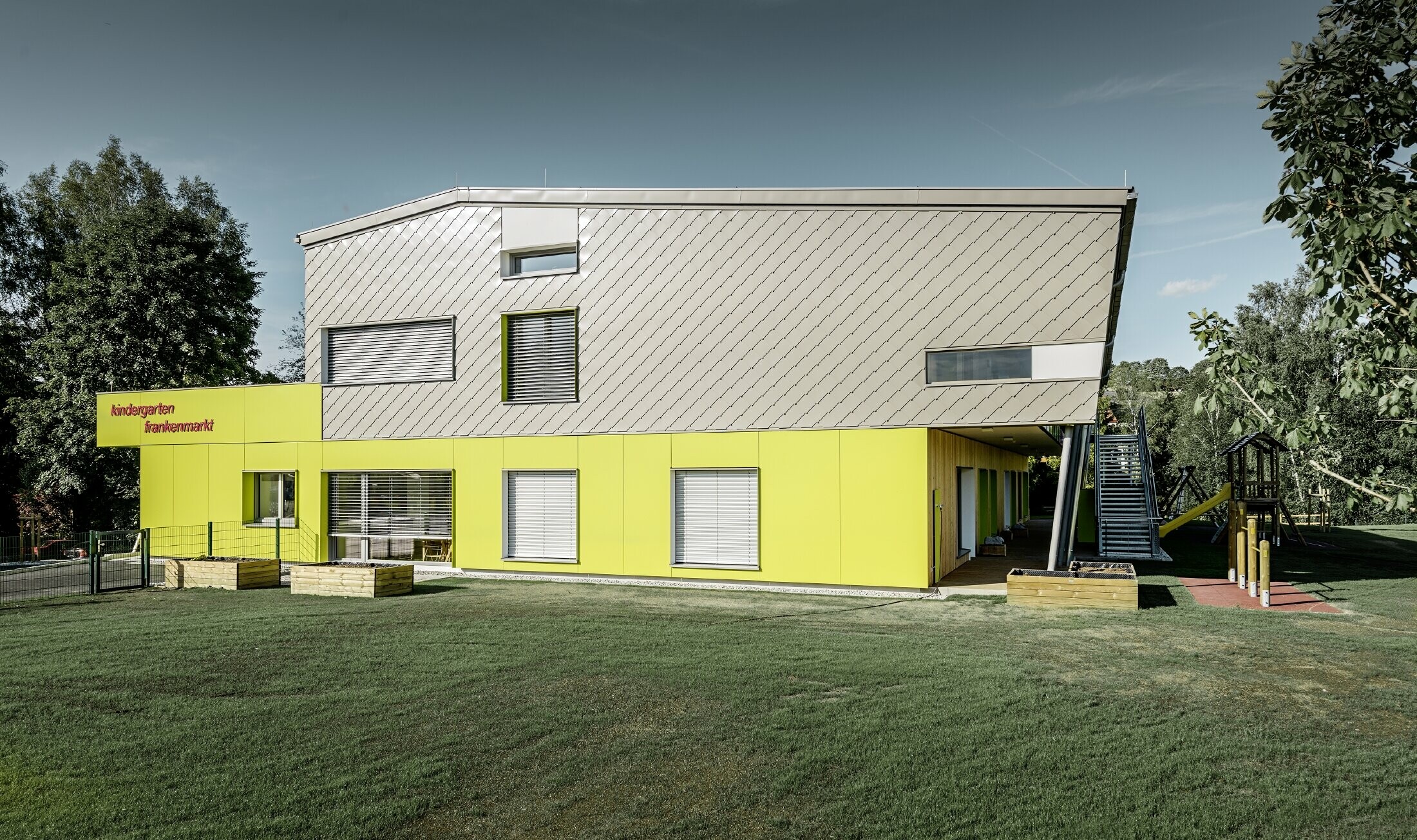 De kleuterschool is deels bekleed met PREFA gevellosange 29x29 in brons, dit gebouwgedeelte heeft ook geen dakoverstek. De rest van de gevel is bekleed met gevelplaten in geel.