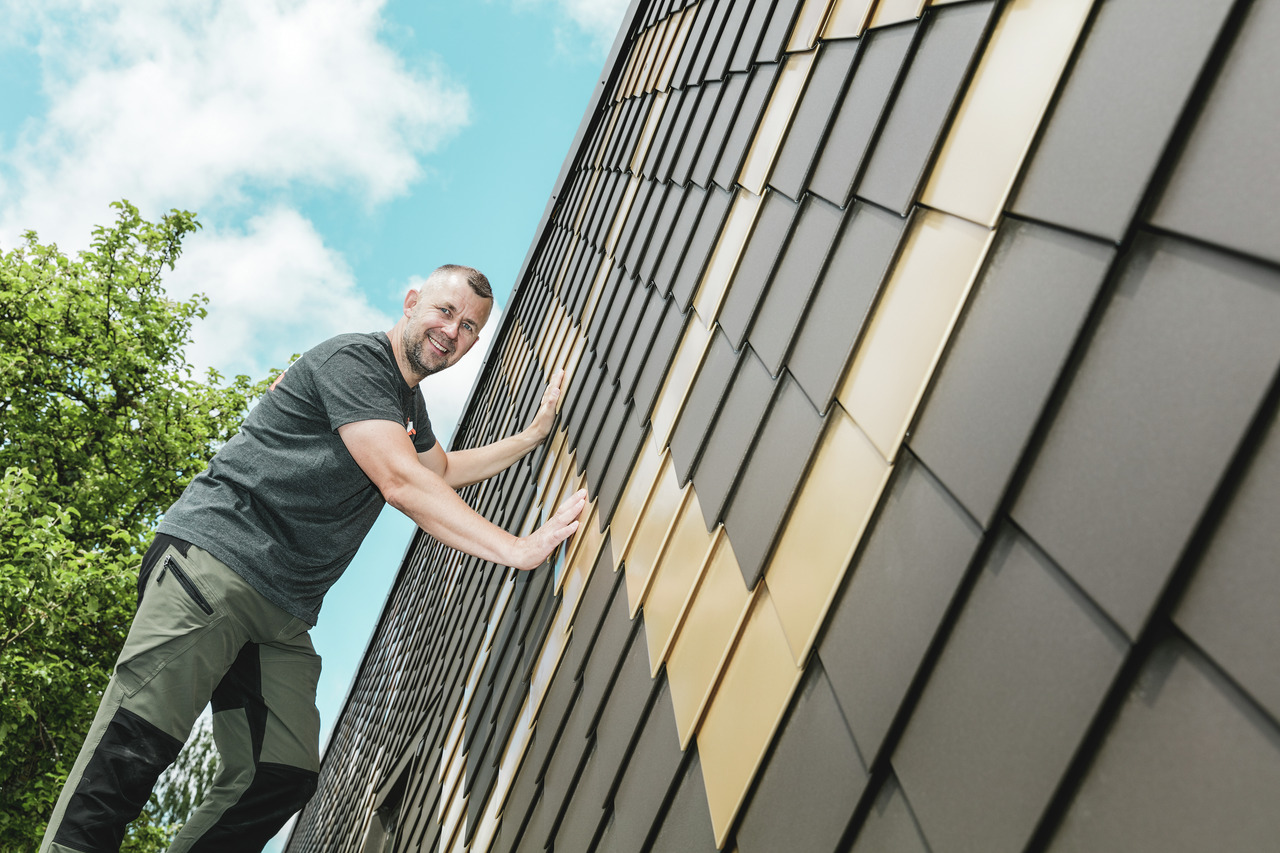 PREFA Handwerker mit grauem T-Shirt an einer PREFA Rauten-Fassade in Braun und Mayagold.